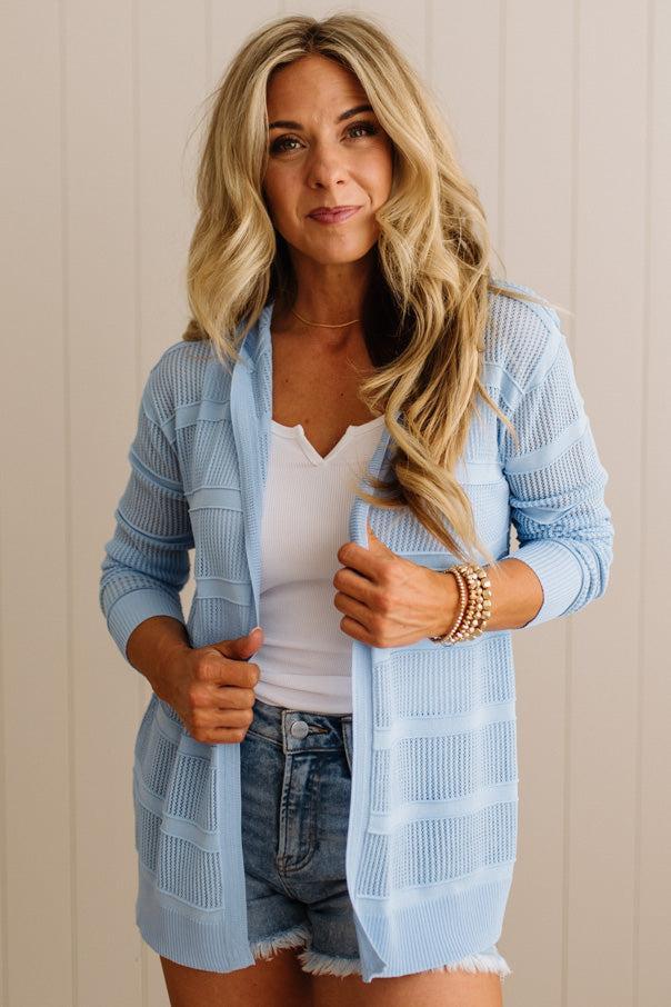 Woman wearing a light blue cardigan over a white top and denim shorts against a beige wall.