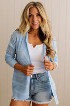 Woman wearing a light blue cardigan, white top, and denim shorts against a beige wall.