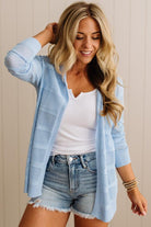 Woman wearing a light blue cardigan over a white top and denim shorts, standing against a neutral background.