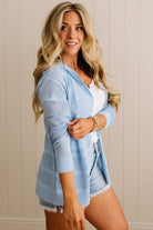 Woman wearing a light blue cardigan over a white top and denim shorts against a beige wall.