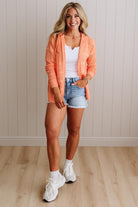 Woman wearing an orange jacket, white top, denim shorts, and white sneakers against a neutral wall.