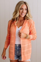 Woman wearing an orange cardigan over a white top and denim shorts, standing against a light background.