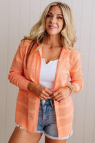 Woman wearing an orange cardigan over a white top and denim shorts against a light background