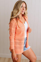 Woman wearing an orange cardigan over a white top and denim shorts indoors.