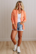 Woman wearing an orange plaid shirt, white top, denim shorts, and white sneakers in a room with wooden flooring and a light-colored wall.