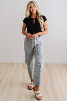 Woman wearing a black top and patterned pants standing in a room with wooden flooring and white walls.