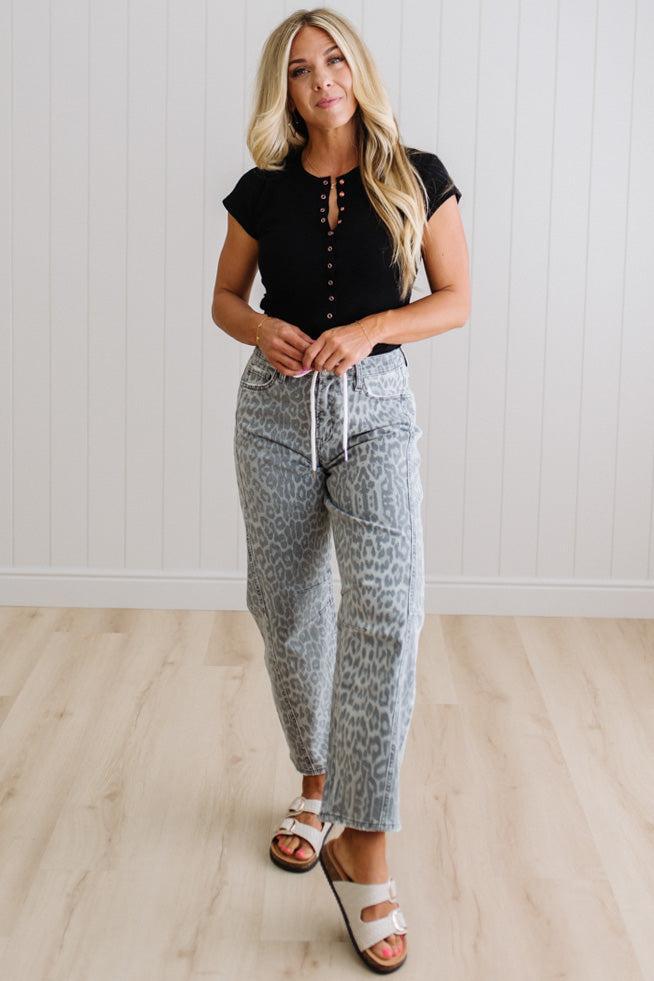 Woman wearing a black top and patterned pants standing in a room with wooden flooring and white walls.