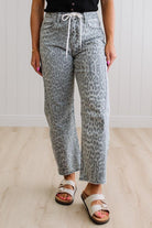 Person wearing leopard print pants and white sandals on a light wooden floor.