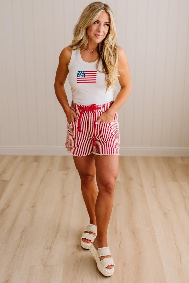 Woman wearing a white tank top with an American flag design and red and white striped shorts on a wooden floor.