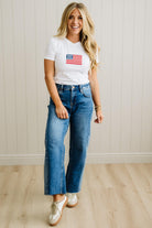 Woman wearing a white t-shirt with an American flag design and blue jeans, standing against a light wooden paneled wall.