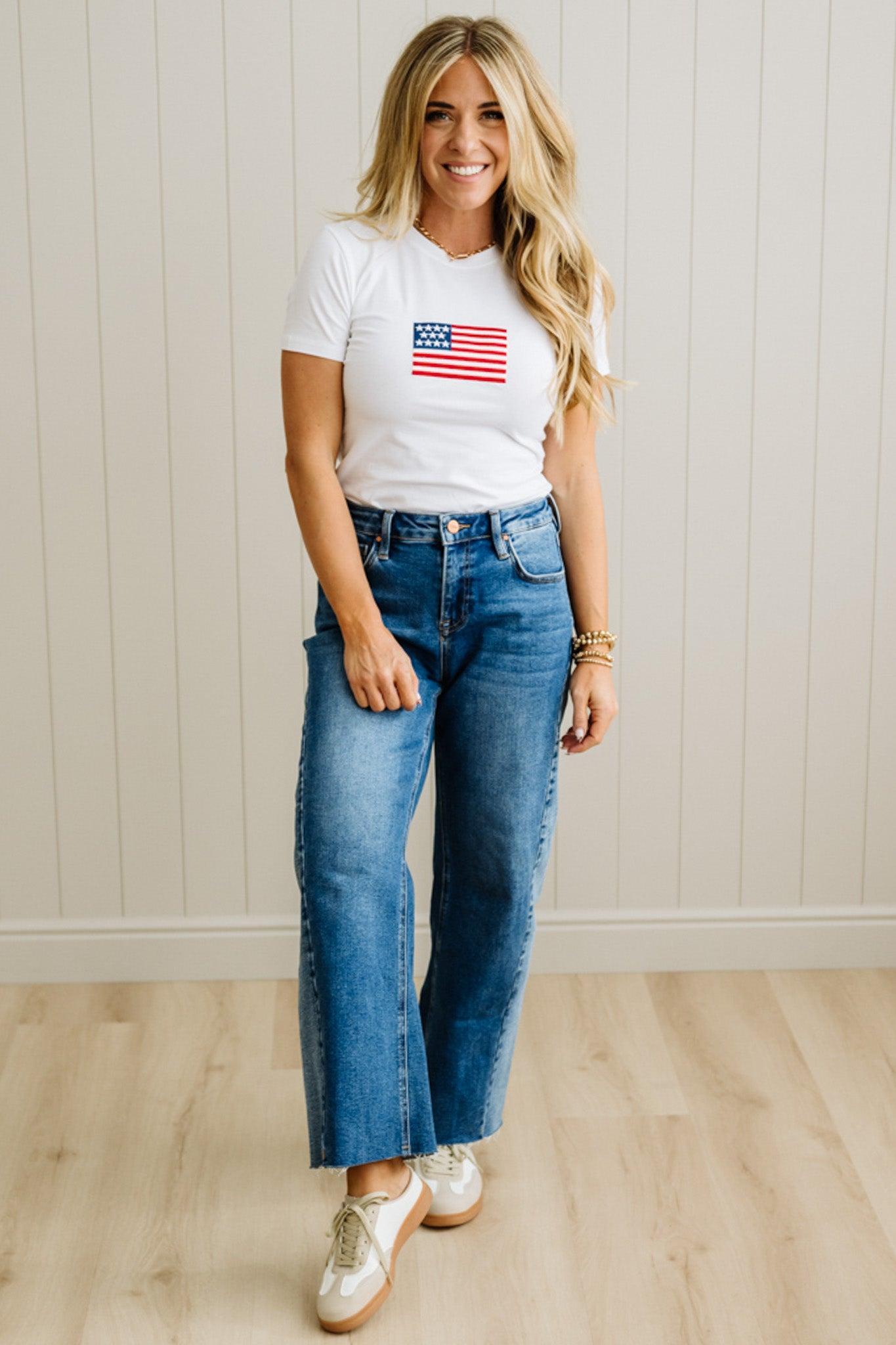 Woman wearing a white t-shirt with an American flag design and blue jeans, standing against a light wooden paneled wall.