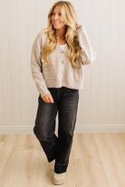 Woman wearing a beige cardigan and dark jeans standing against a white wall.