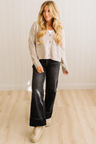 Woman wearing a beige cardigan, white shirt, black flared jeans, and beige shoes in a room with wooden flooring and a white wall.