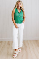 Woman wearing a green sleeveless top and white pants standing against a light wooden paneled wall.