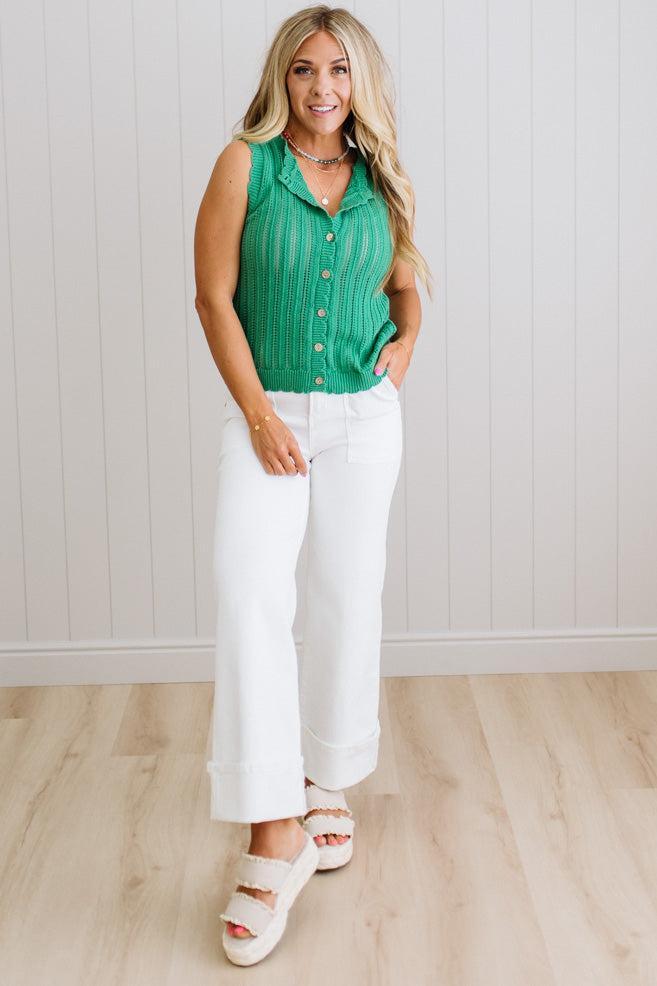 Woman wearing a green sleeveless top and white pants standing against a light wooden paneled wall.