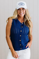 Woman wearing a blue sleeveless cardigan and white cap with a logo, standing against a plain background.