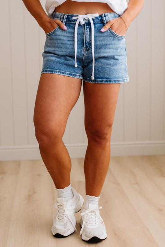 Person wearing denim shorts and white sneakers in a room with wooden floor and light-colored wall.
