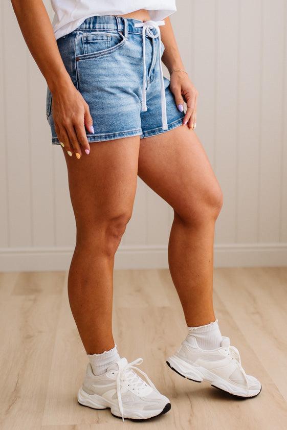 Person wearing denim shorts and white sneakers on a wooden floor.