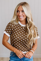 A smiling blonde woman wearing a brown short-sleeve top with white polka dots and white ruffled trim, paired with blue jeans, standing against a light paneled wall.