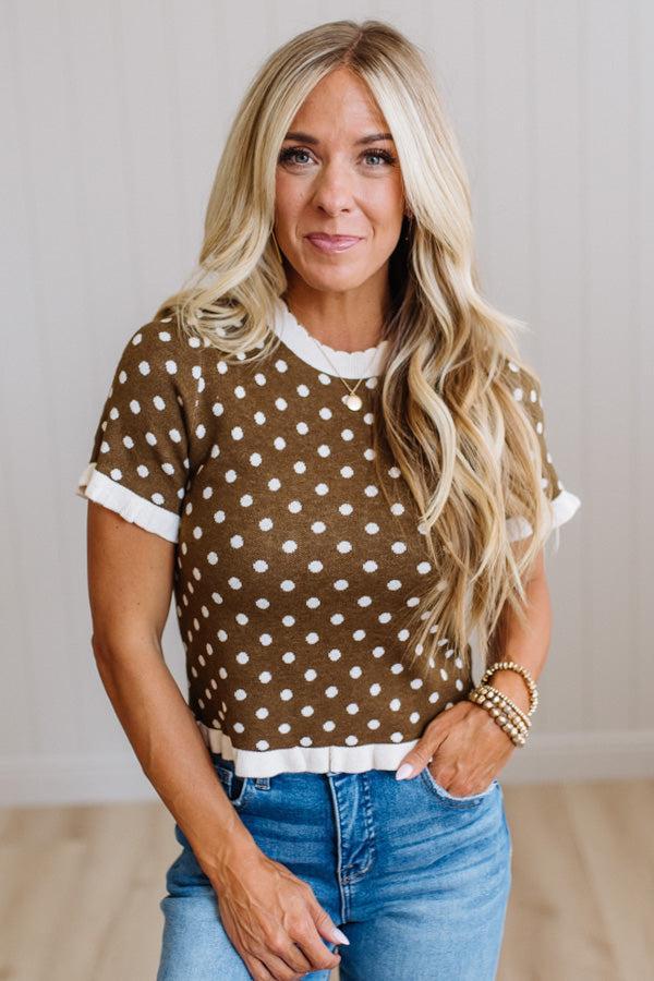 A woman with long blonde hair wearing a brown short-sleeve knit top with white polka dots and white ruffled trim, paired with blue jeans, standing against a light-colored paneled wall.