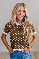 A smiling blonde woman wearing a brown short-sleeve top with white polka dots and white ruffled trim, paired with blue jeans, standing against a light neutral paneled wall.