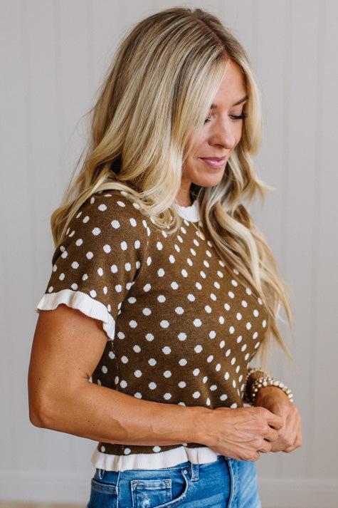 A woman wearing a brown short-sleeve knit top with white polka dots and white ruffled trim on the sleeves, paired with blue jeans.