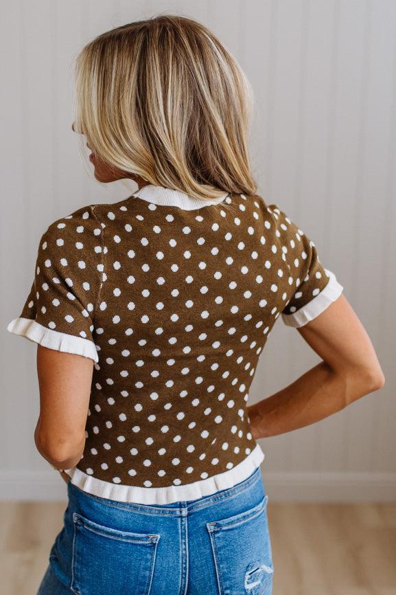 A woman shown from the back wearing a brown short-sleeve top with white polka dots and white ruffled trim, paired with blue jeans, in an indoor setting.
