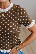 A close-up of a woman wearing a brown short-sleeve knit top with white polka dots and white ruffled trim at the collar, sleeve cuffs, and hem, paired with blue jeans and layered pearl bracelets.