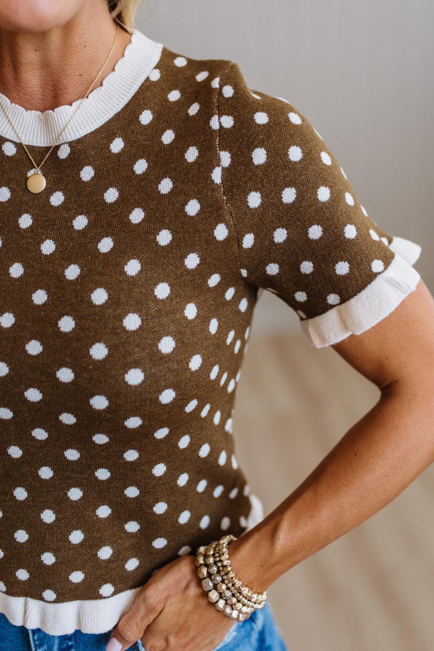 A close-up of a woman wearing a brown short-sleeve knit top with white polka dots and white ruffled trim at the collar, sleeve cuffs, and hem, paired with blue jeans and layered pearl bracelets.