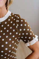 Close-up of a woman wearing a brown short-sleeve knit top with white polka dots and white ruffled trim at the collar and sleeve cuffs.