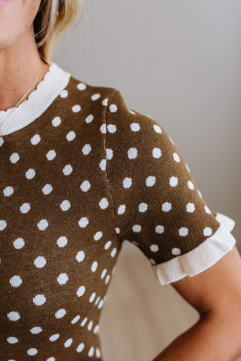 Close-up of a woman wearing a brown short-sleeve knit top with white polka dots and white ruffled trim at the collar and sleeve cuffs.
