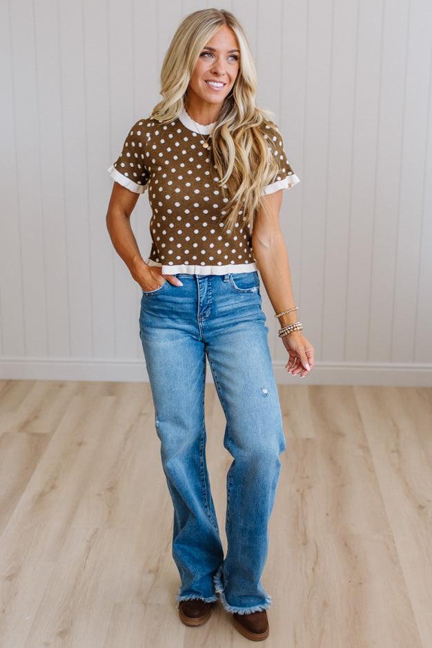 A woman modeling a brown short-sleeve top with white polka dots and white ruffle trim, paired with blue jeans and brown shoes, standing in a room with light wood floors and white paneled walls.