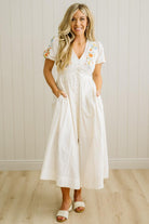 Woman wearing a white dress with floral embroidery against a light wooden paneled wall.