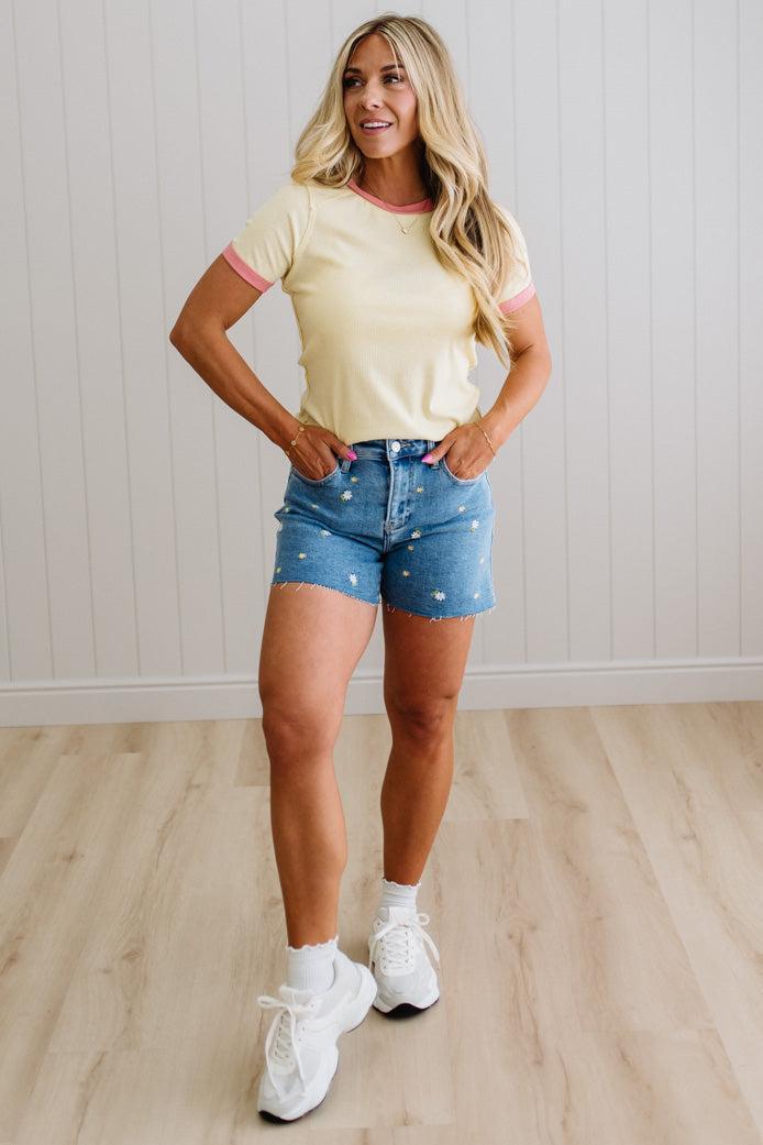 Woman wearing a yellow t-shirt and denim shorts standing on a wooden floor with a white paneled wall background.