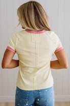 Person wearing a light yellow shirt with pink trim and blue jeans, standing against a plain background