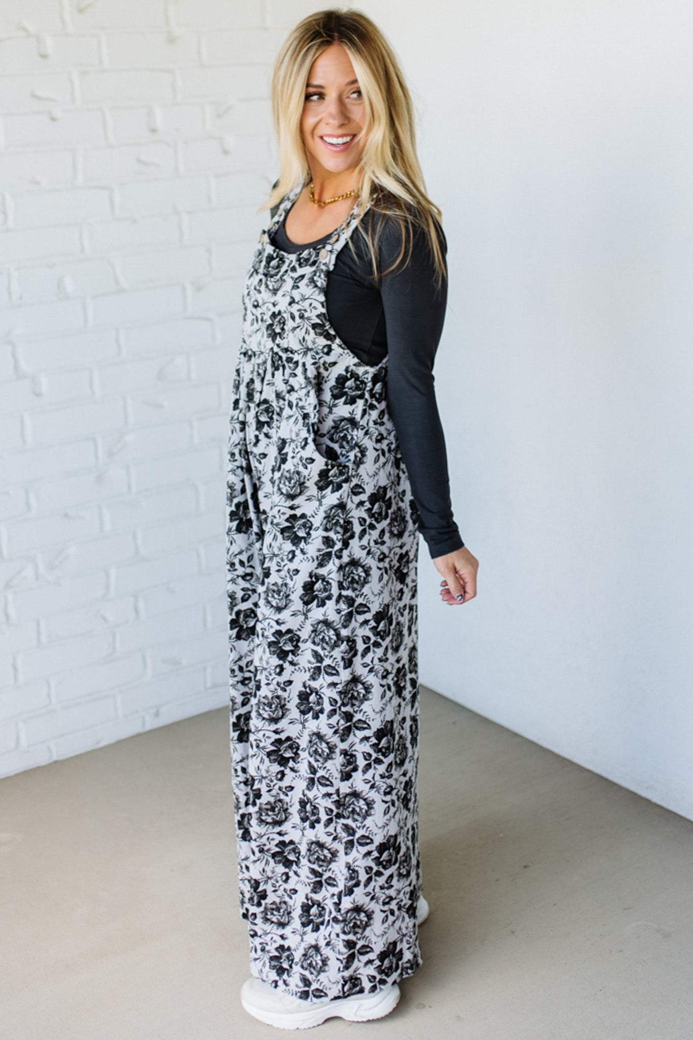 Woman wearing a black floral dress over a black long-sleeve shirt against a white brick wall.