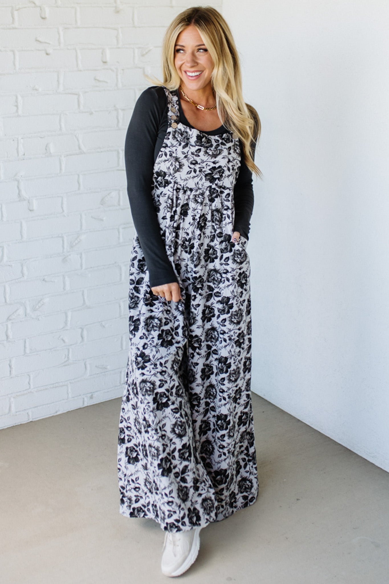 Woman wearing a floral overalls dress against a white brick wall.