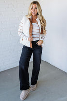 Woman wearing a striped shirt, white jacket, and dark jeans against a white brick wall.