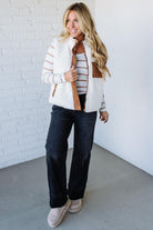 Woman wearing a white and brown striped jacket with black pants against a white brick wall.