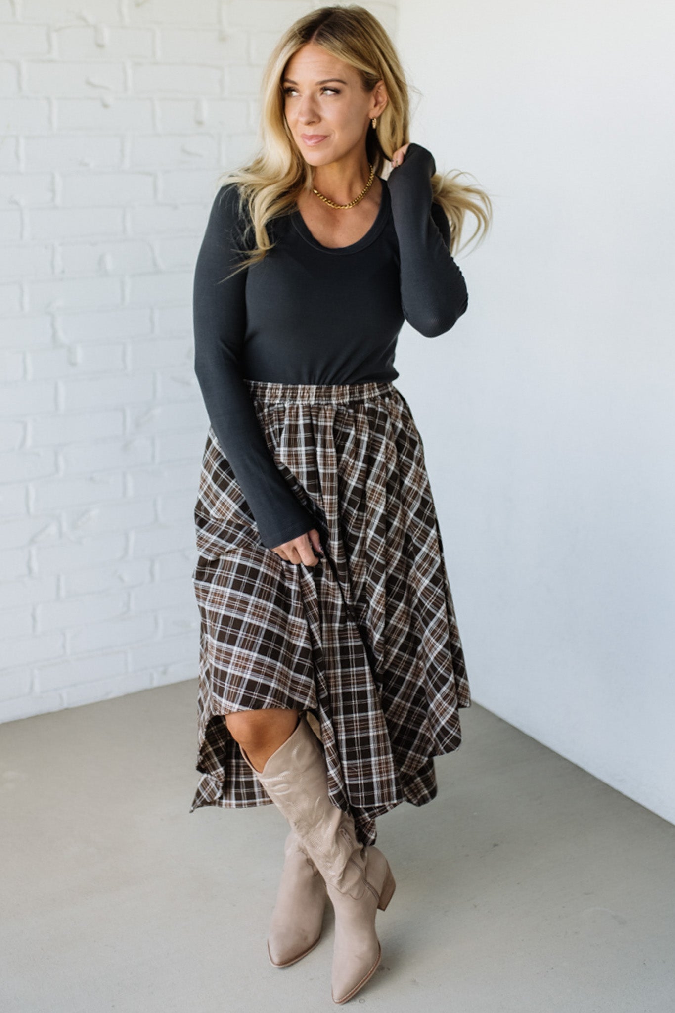 Woman wearing a black long-sleeve top and plaid skirt with beige knee-high boots against a white brick wall.