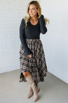 Woman wearing a black long-sleeve top and plaid skirt with beige knee-high boots against a white brick wall.