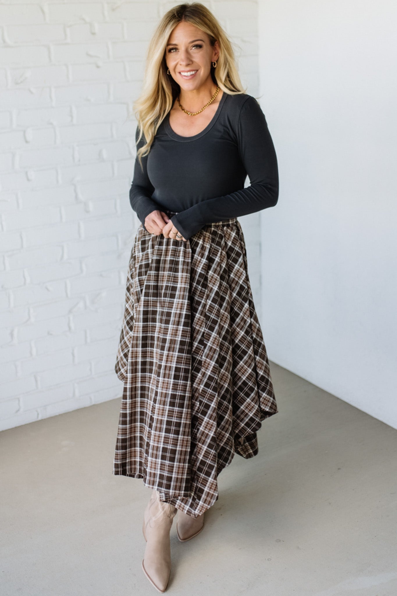 Woman wearing a black long-sleeve top and plaid skirt against a white brick wall.