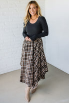 Woman wearing a black long-sleeve top and plaid skirt against a white brick wall.