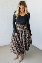 Woman wearing a black long-sleeve top and plaid skirt against a white brick wall.