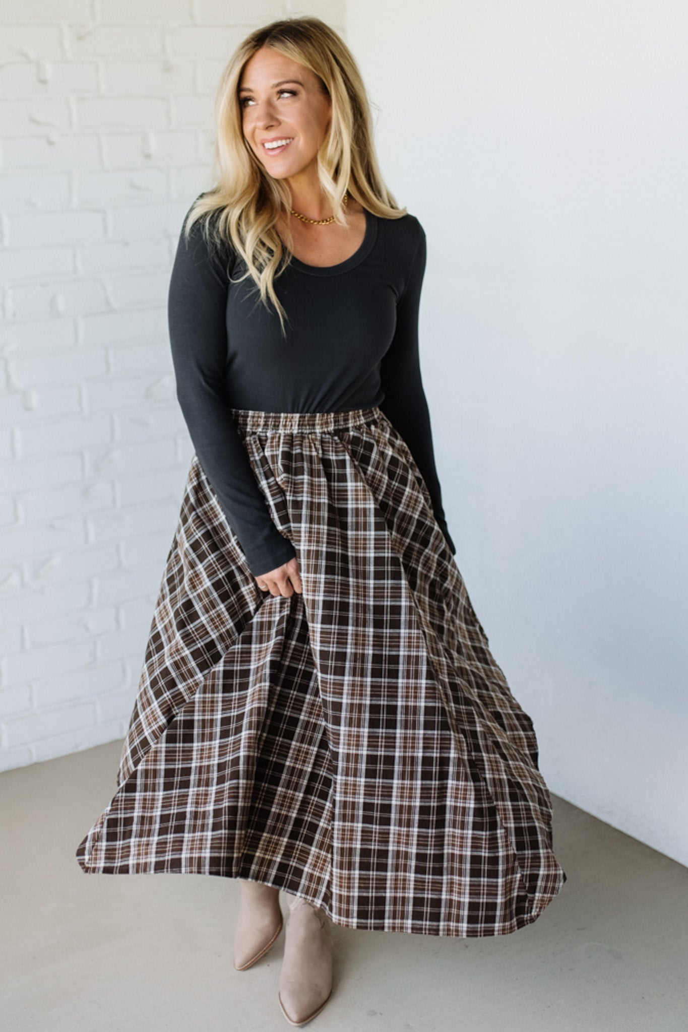 Woman wearing a black top and plaid skirt against a white brick wall.