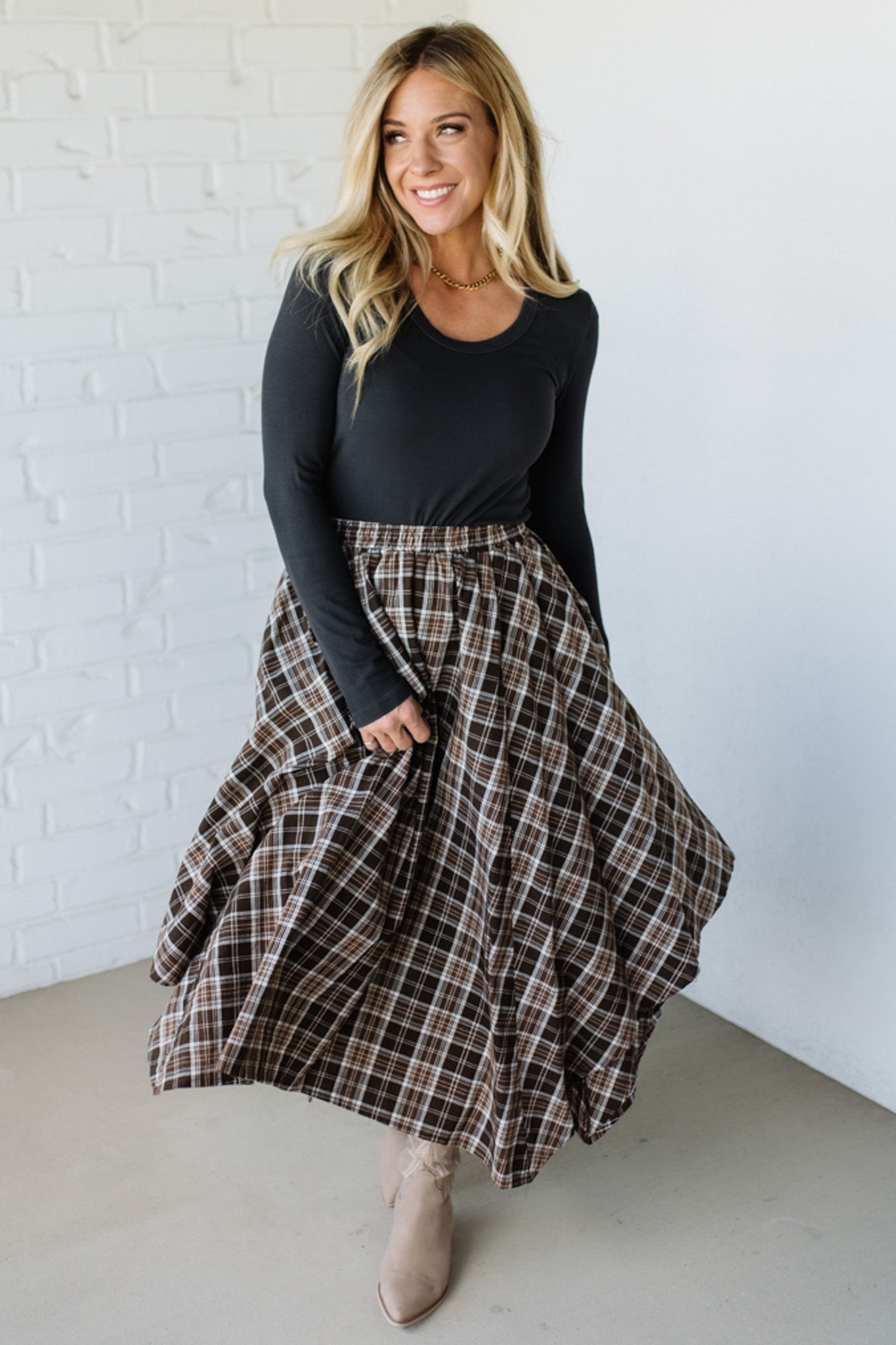 Woman wearing a black long-sleeve top and plaid skirt against a white brick wall.
