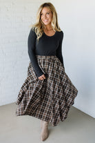 Woman wearing a black long-sleeve top and plaid skirt against a white brick wall.