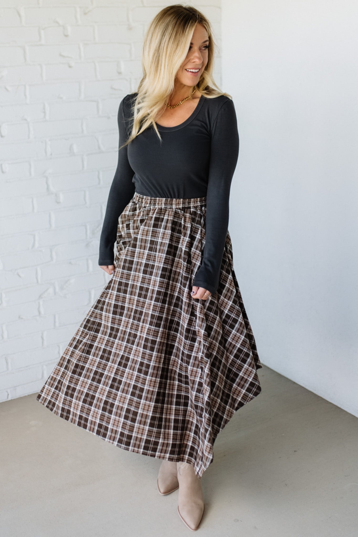 Woman wearing a black long-sleeve top and plaid skirt against a white brick wall.