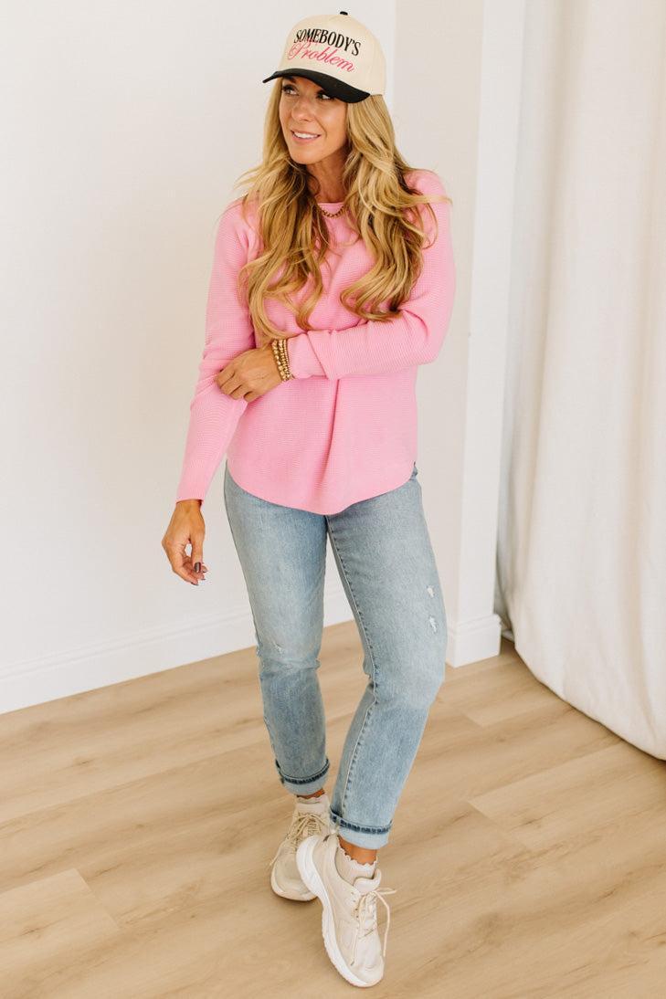 Blonde woman wearing jeans and a pink sweater with a black and white hat and sneakers.