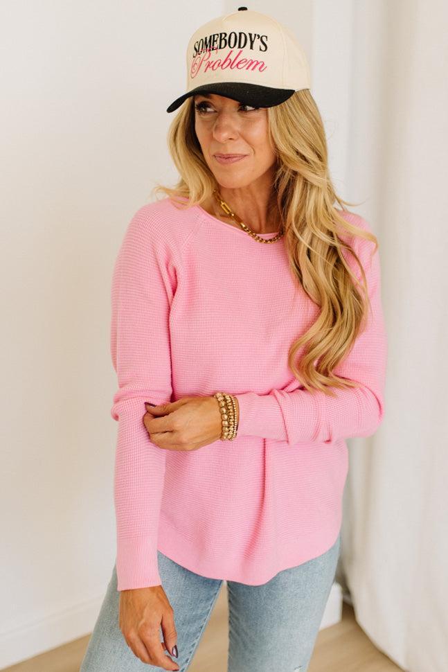 Blonde woman wearing jeans and a pink sweater with a black and white hat and sneakers.